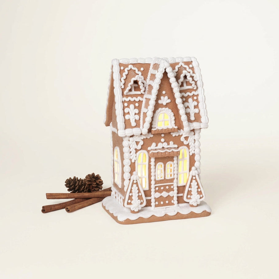 A gingerbread house-shaped decorative home figure, dough, with white icing-style detailing and warm yellow lights glowing from within, shown on a plain beige background with cinnamon sticks and a pinecone beside it.