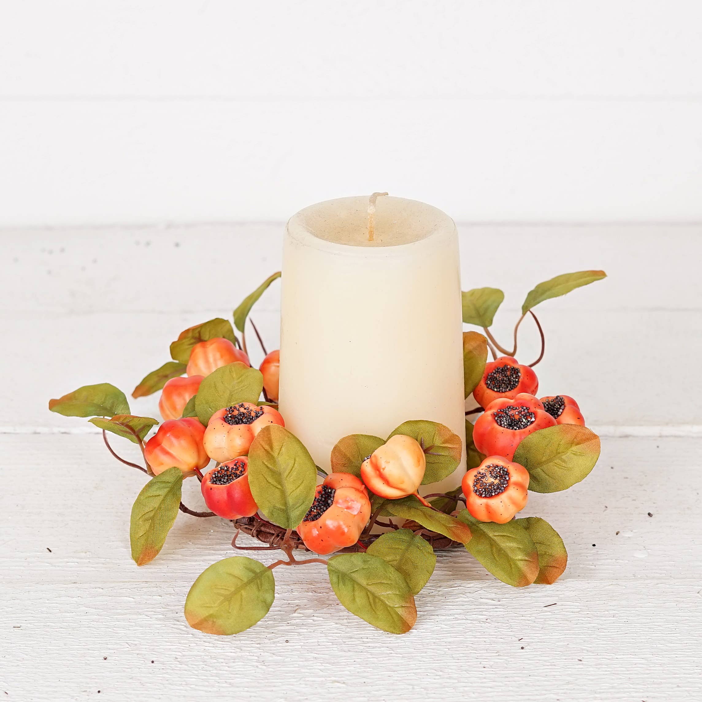 An off-white pillar candle centered in a decorative wreath made of green leaves and orange-red berries with dark seeds, displayed on a white painted wood surface against a white background.