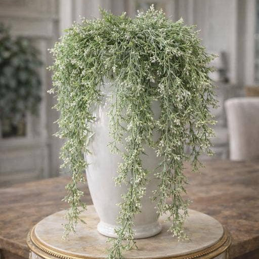 A large white ceramic urn filled with cascading green and white variegated foliage, displayed on an ornate round table in a softly blurred interior setting.