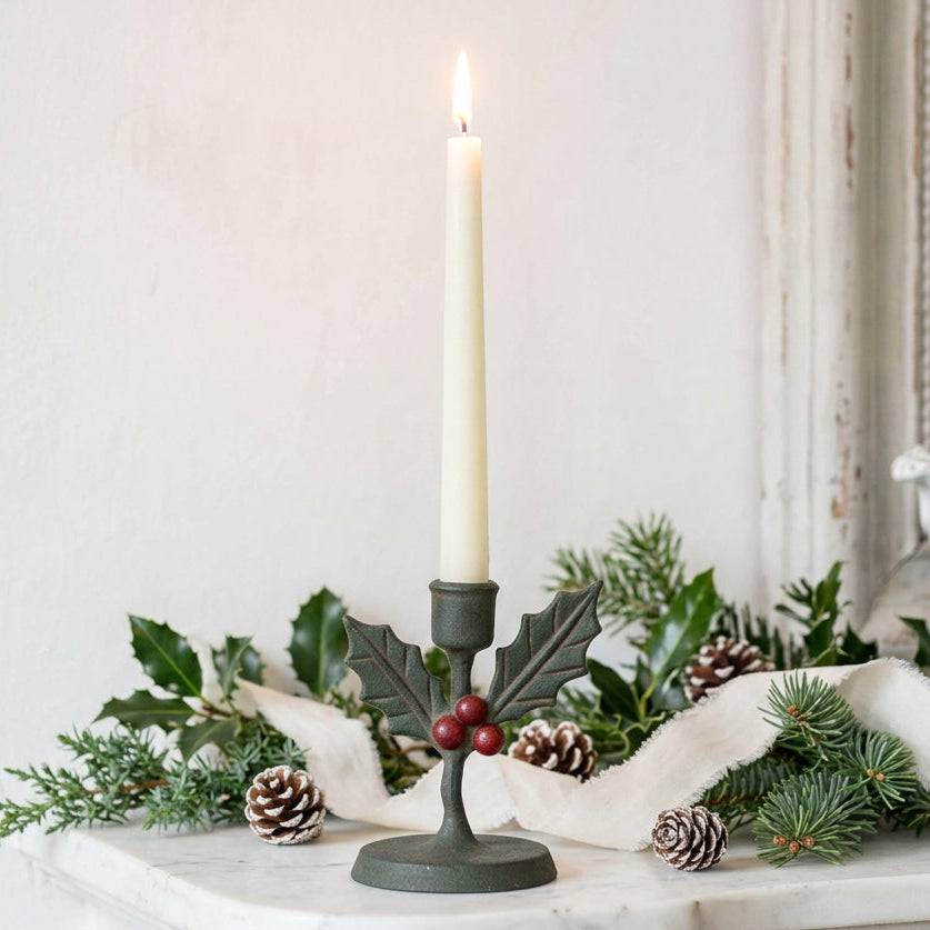 A dark green metal candle holder shaped like holly with red berries, holding a lit white taper candle, set on a rustic wooden surface against a plain beige wall.