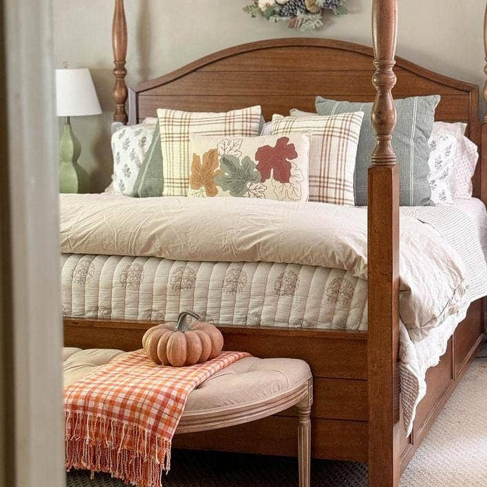 A warm wood four-poster bed with a quilted cream bedding set, styled with patterned pillows, a matching bench with a plaid throw, and a small decorative pumpkin in a bedroom setting.