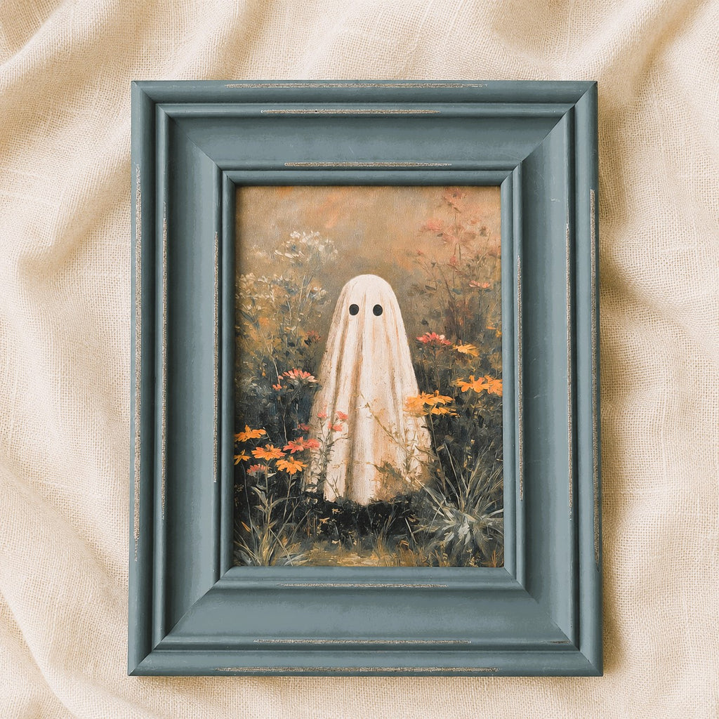 A framed art print featuring a whimsical painting of a ghost in a white sheet standing in a field of orange flowers, set in a distressed dark teal frame on a beige fabric background.