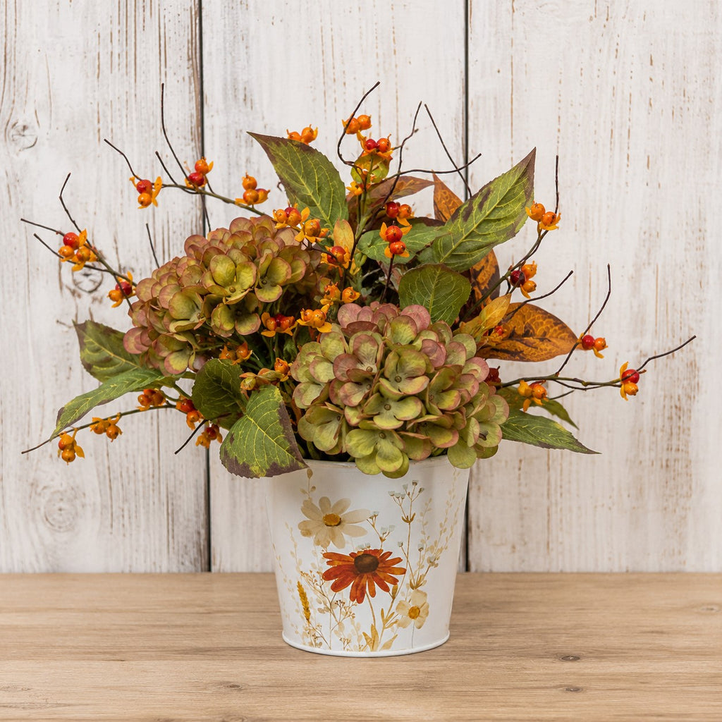 An artificial autumn floral arrangement featuring hydrangea-like blooms in muted green and pinkish tones, orange berries, and brown leaves, displayed in a white bucket with a floral print against a whitewashed wood plank background.