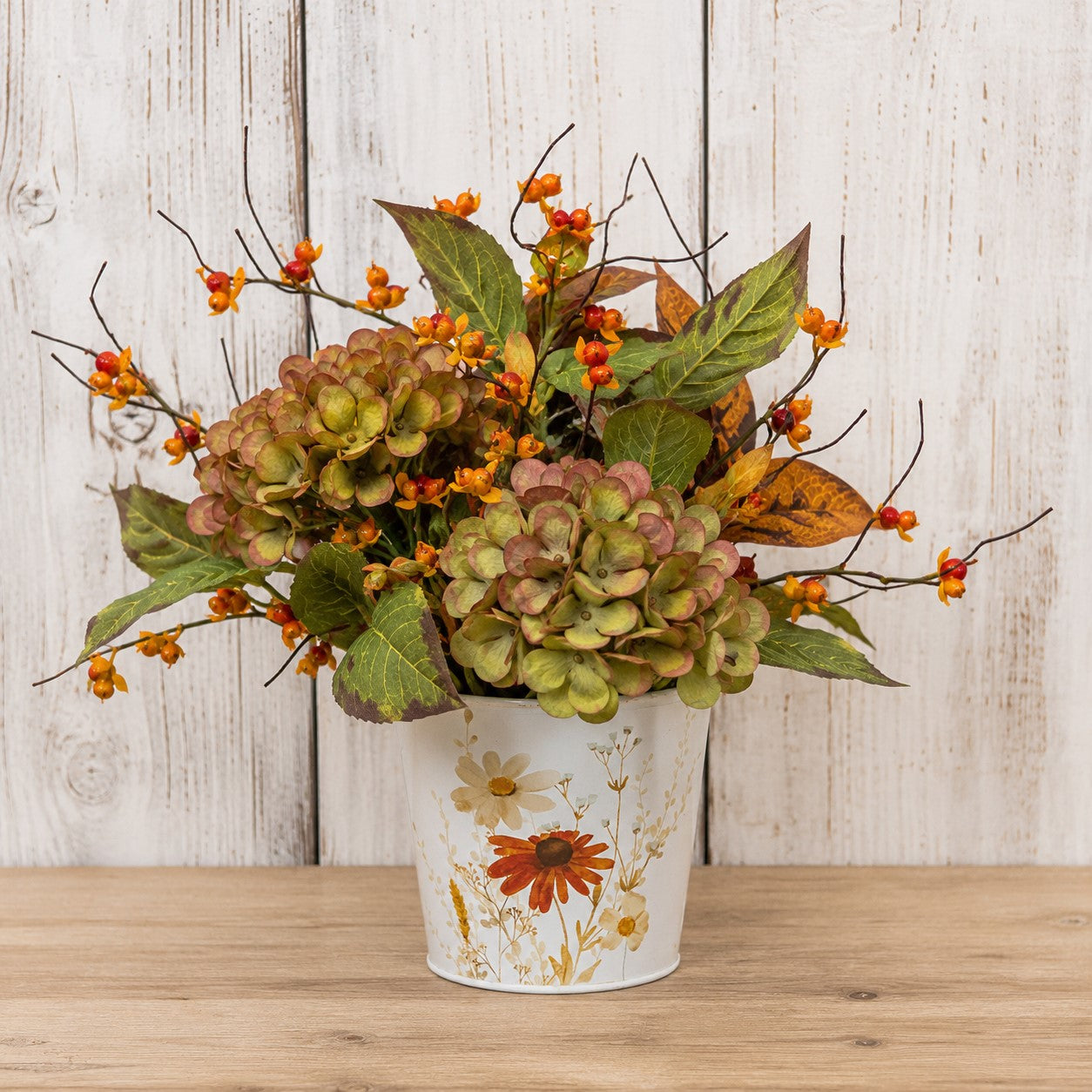 An artificial autumn floral arrangement featuring hydrangea-like blooms in muted green and pinkish tones, orange berries, and brown leaves, displayed in a white bucket with a floral print against a whitewashed wood plank background.