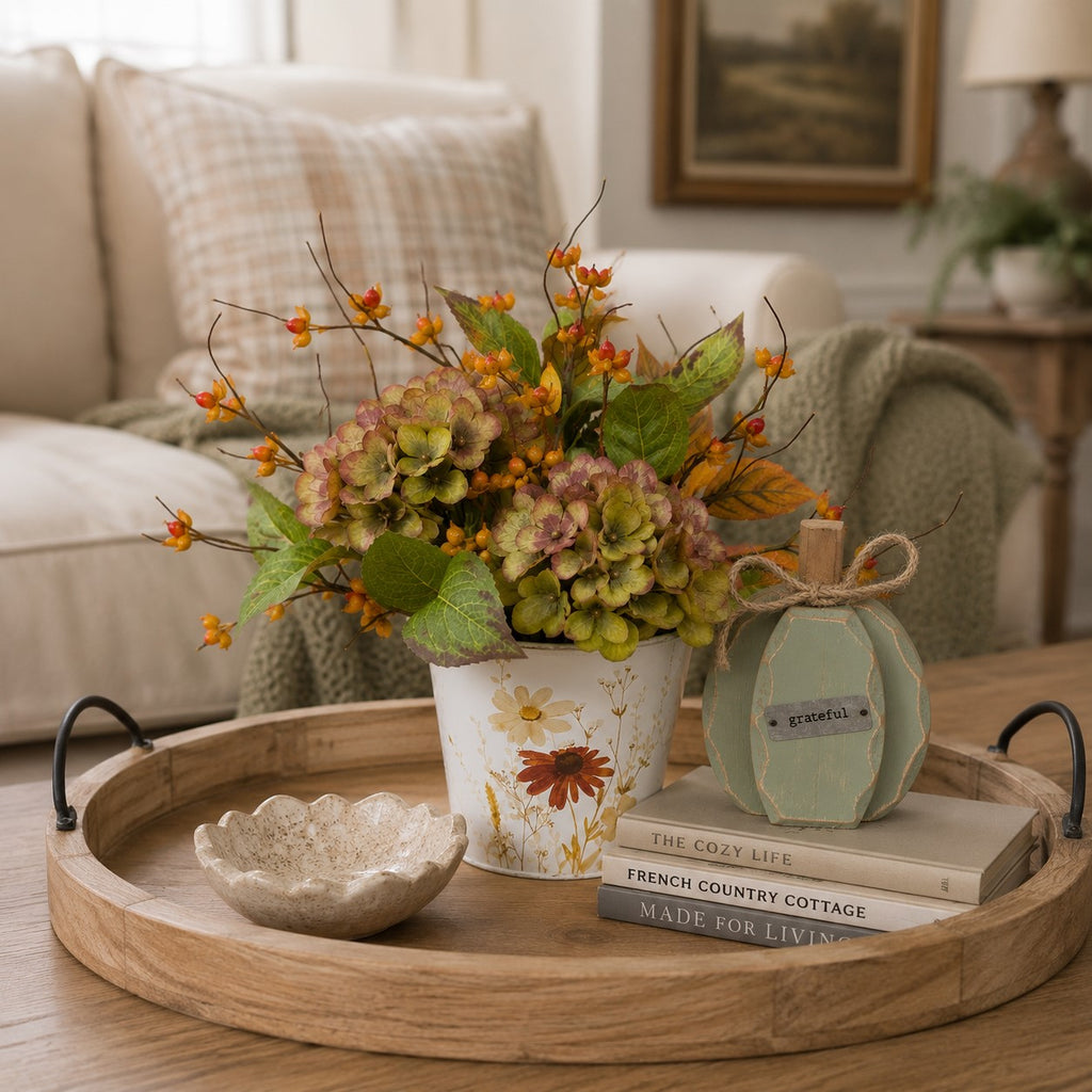 A styled home decor scene on a wooden tray in a living room, featuring a floral-patterned vase with hydrangea-like flowers and orange berries, a pale green decorative pumpkin, and stacked books.
