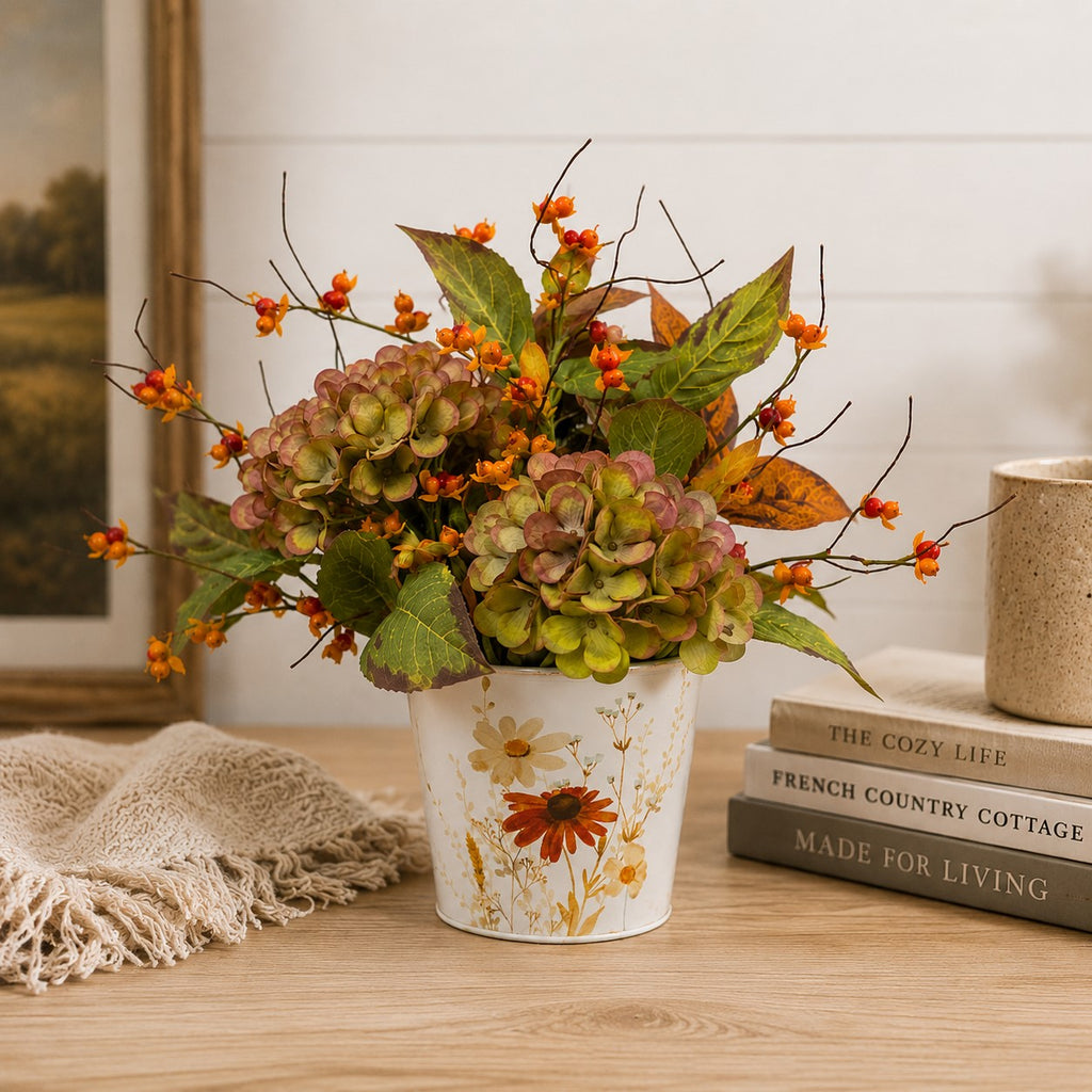 An artificial autumn floral arrangement in a white ceramic pot with orange flowers, green and brown hydrangea-like blooms, and fall leaves, styled on a wooden table with books, a throw blanket, and a mug against a white shiplap wall.