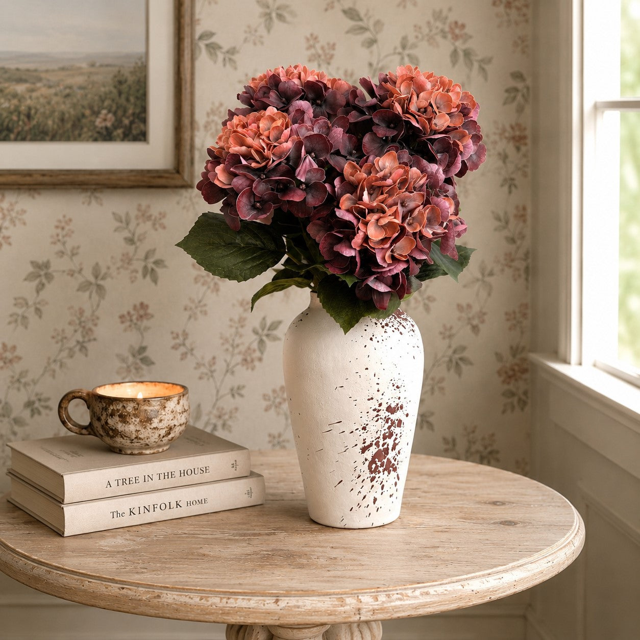 A bouquet of artificial hydrangea flowers in shades of dusty pink, terracotta, and deep burgundy, arranged in a distressed white ceramic vase with brown speckles, set on a round wooden table next to a stack of books and a small cup.
