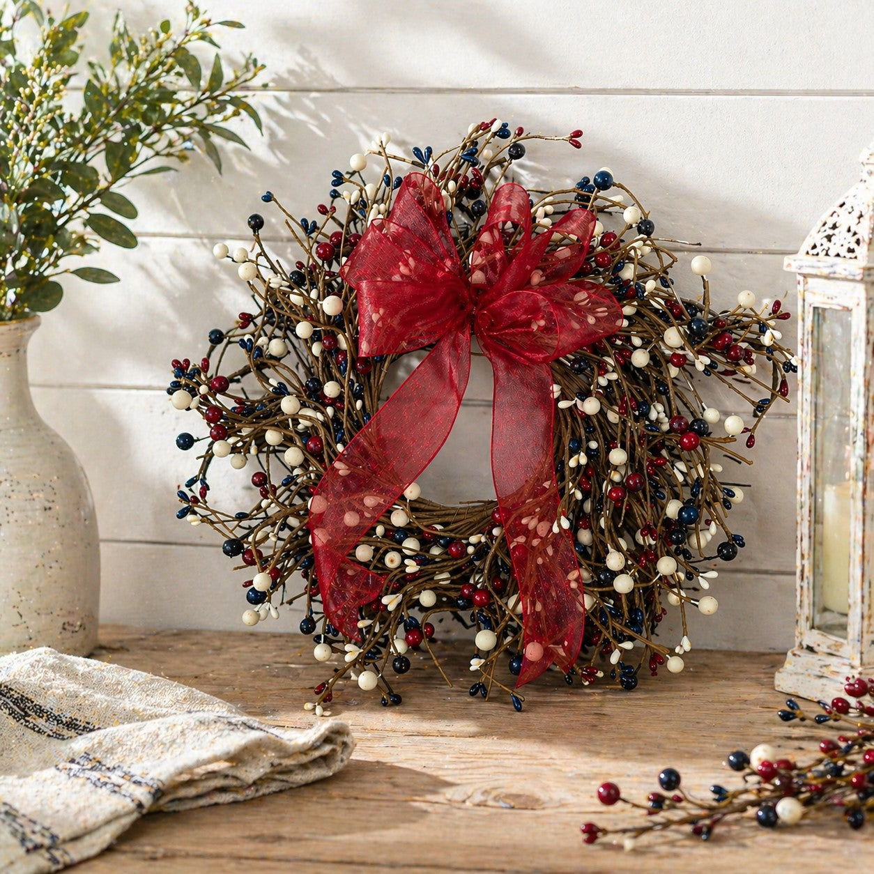 Patriotic berry wreath with red bow on a wooden surface, surrounded by plants and a lantern.