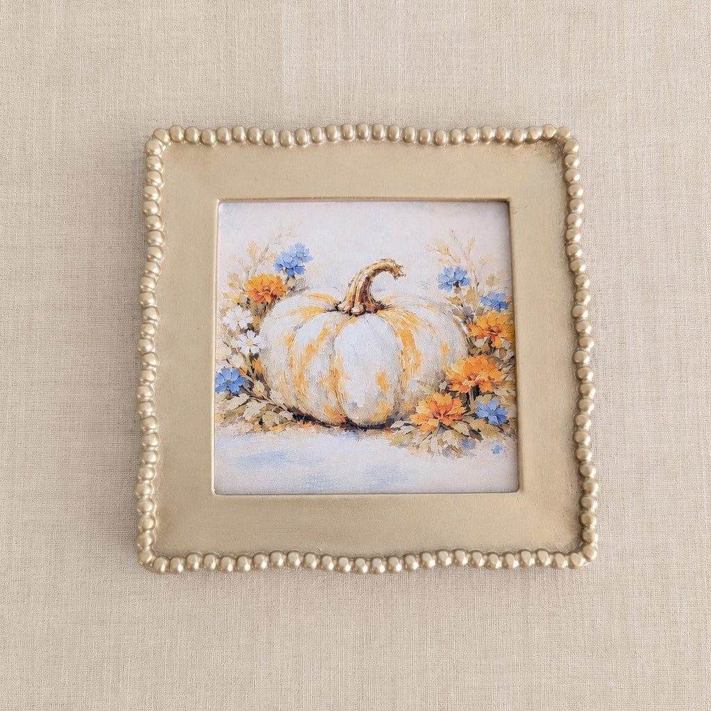 A square decorative picture frame with a beaded border, showcasing a painted illustration of a white pumpkin surrounded by orange and blue flowers, set against a neutral beige textured background.