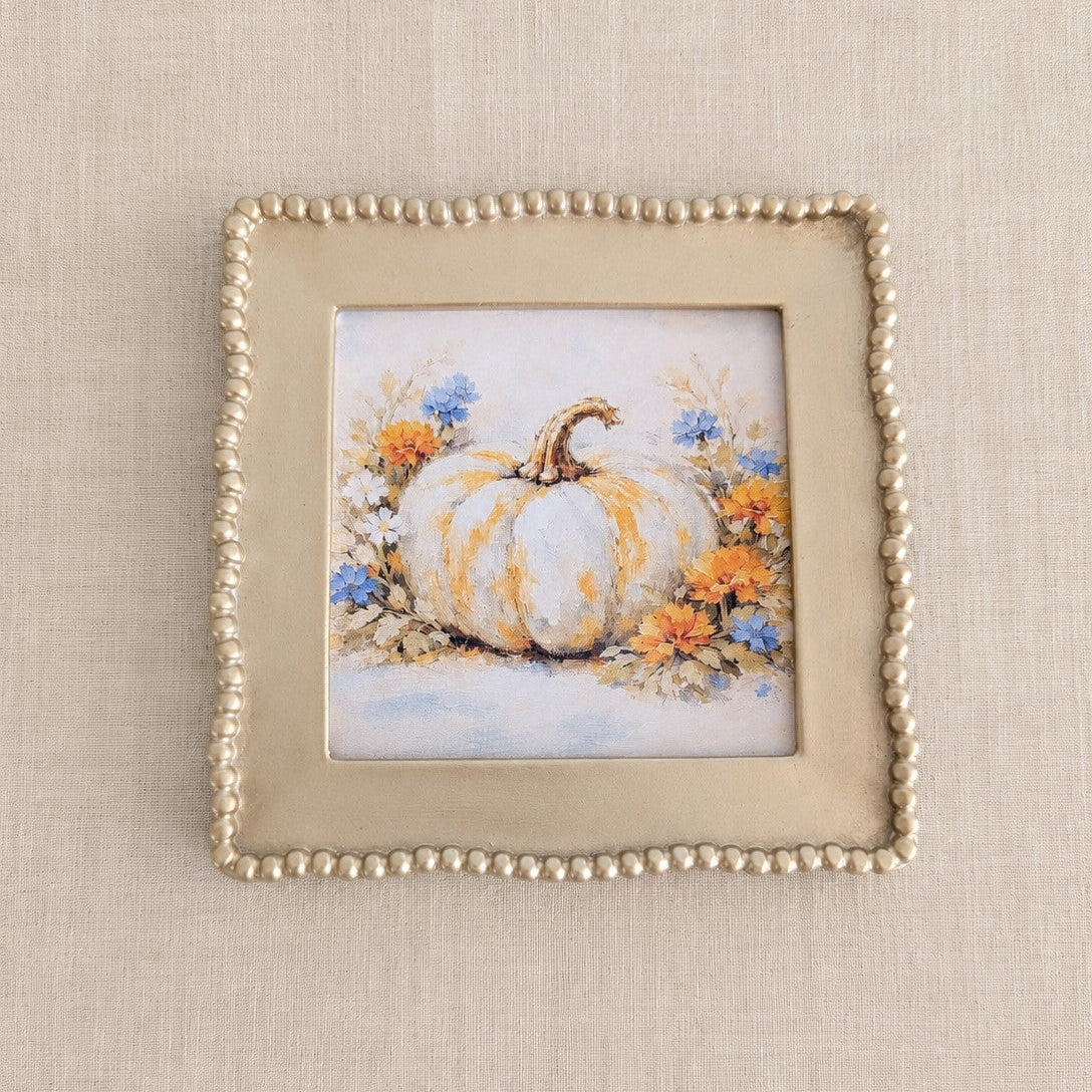 A square decorative picture frame with a beaded border, showcasing a painted illustration of a white pumpkin surrounded by orange and blue flowers, set against a neutral beige textured background.