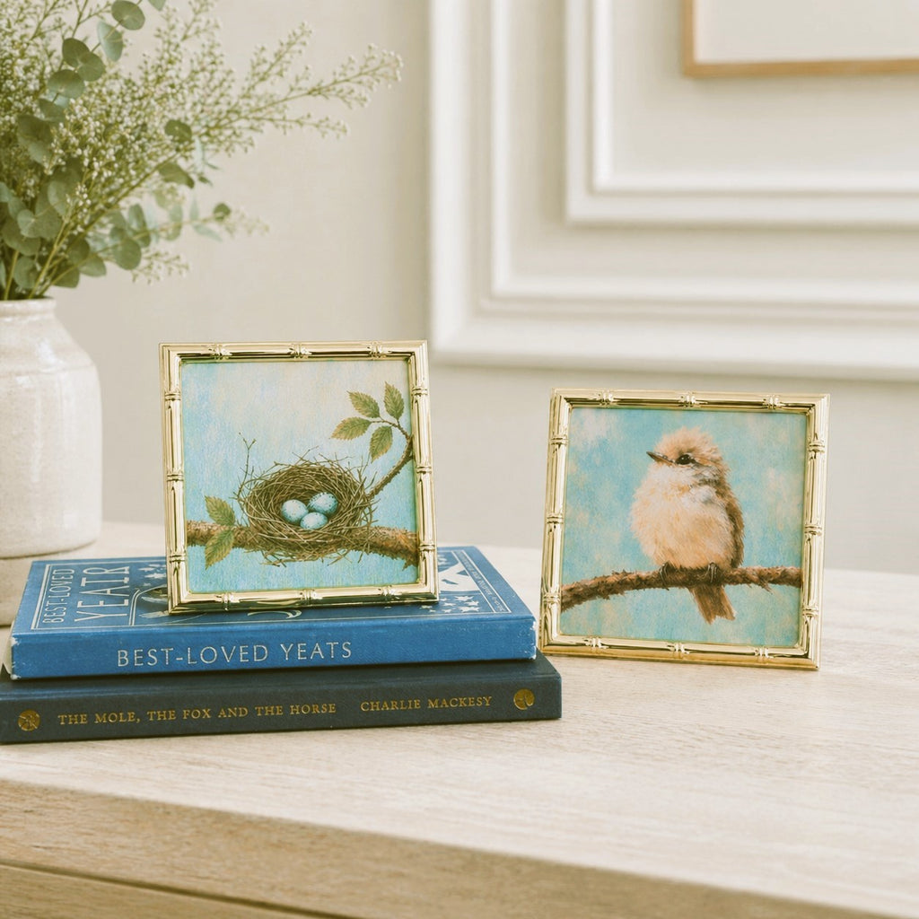 Two framed artworks of birds and a nest on top of books with a vase of flowers in the background.