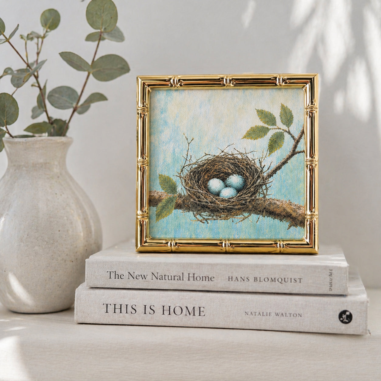 Decorative framed print of a nest with eggs on books, next to a vase with greenery.