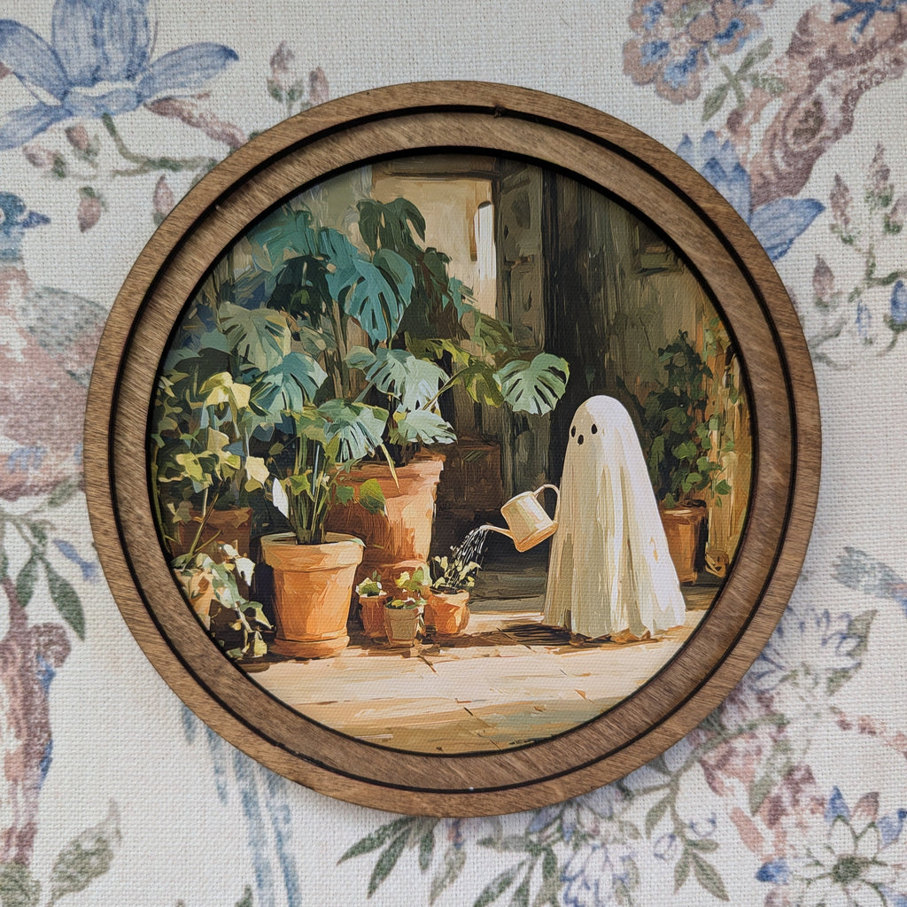 Round framed wall art featuring a whimsical painting of a ghost in a white sheet watering potted plants, mounted on a floral wallpaper background.