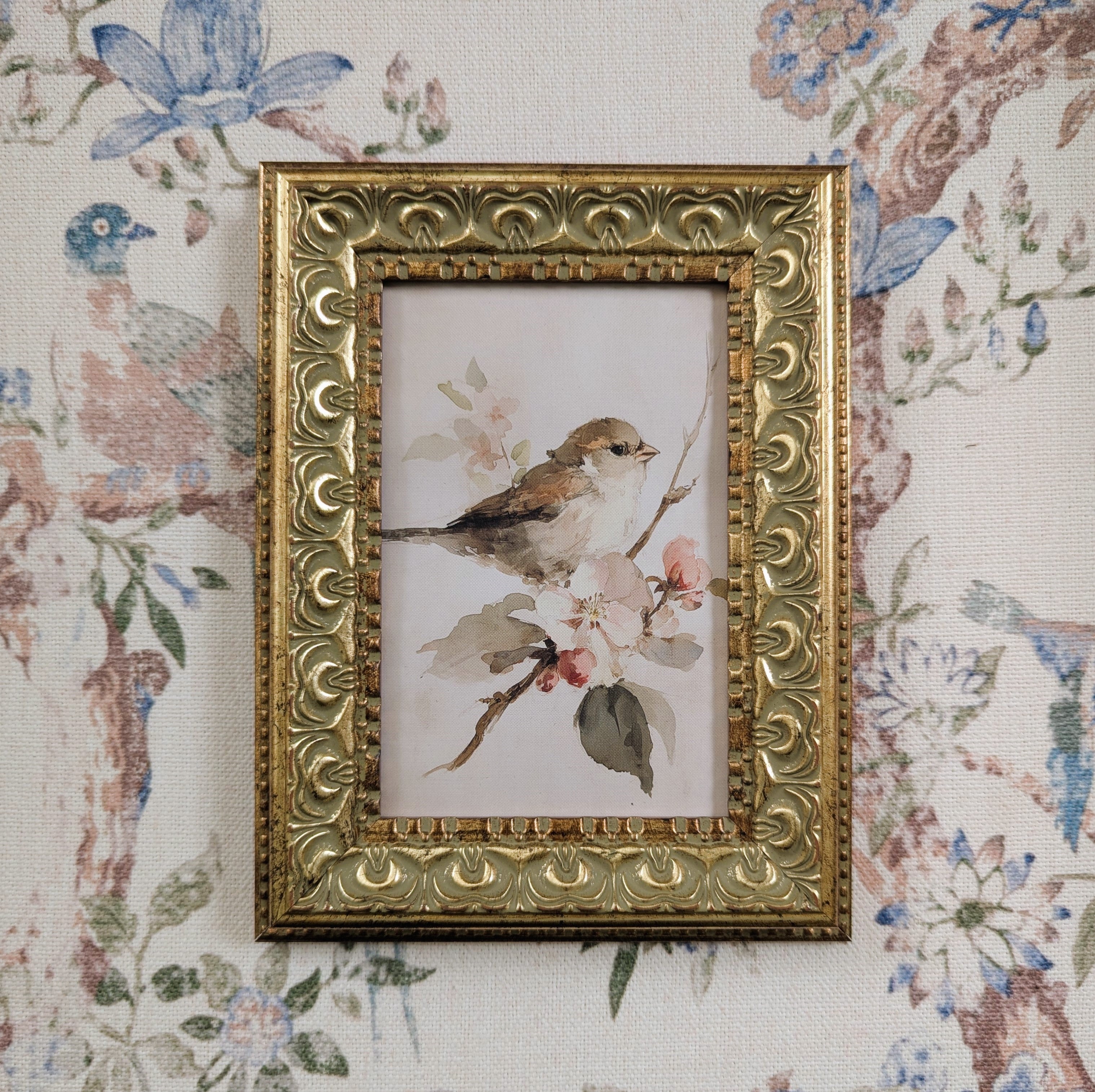 A small ornate gold picture frame holding a watercolor-style bird perched among pink blossoms, displayed against a floral wallpaper background.