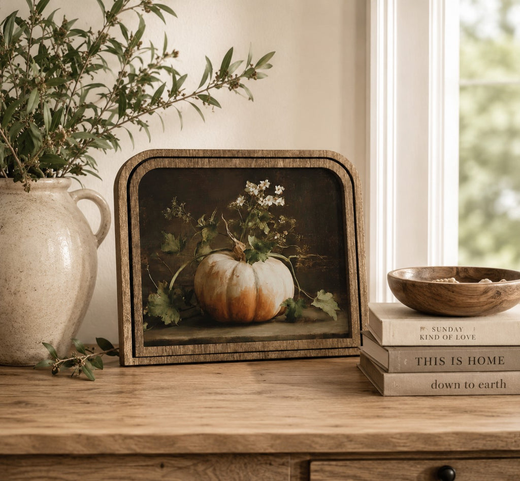 A framed still life art print featuring a small orange pumpkin and white flowers, styled on a wooden table next to a ceramic vase with greenery and a stack of books.