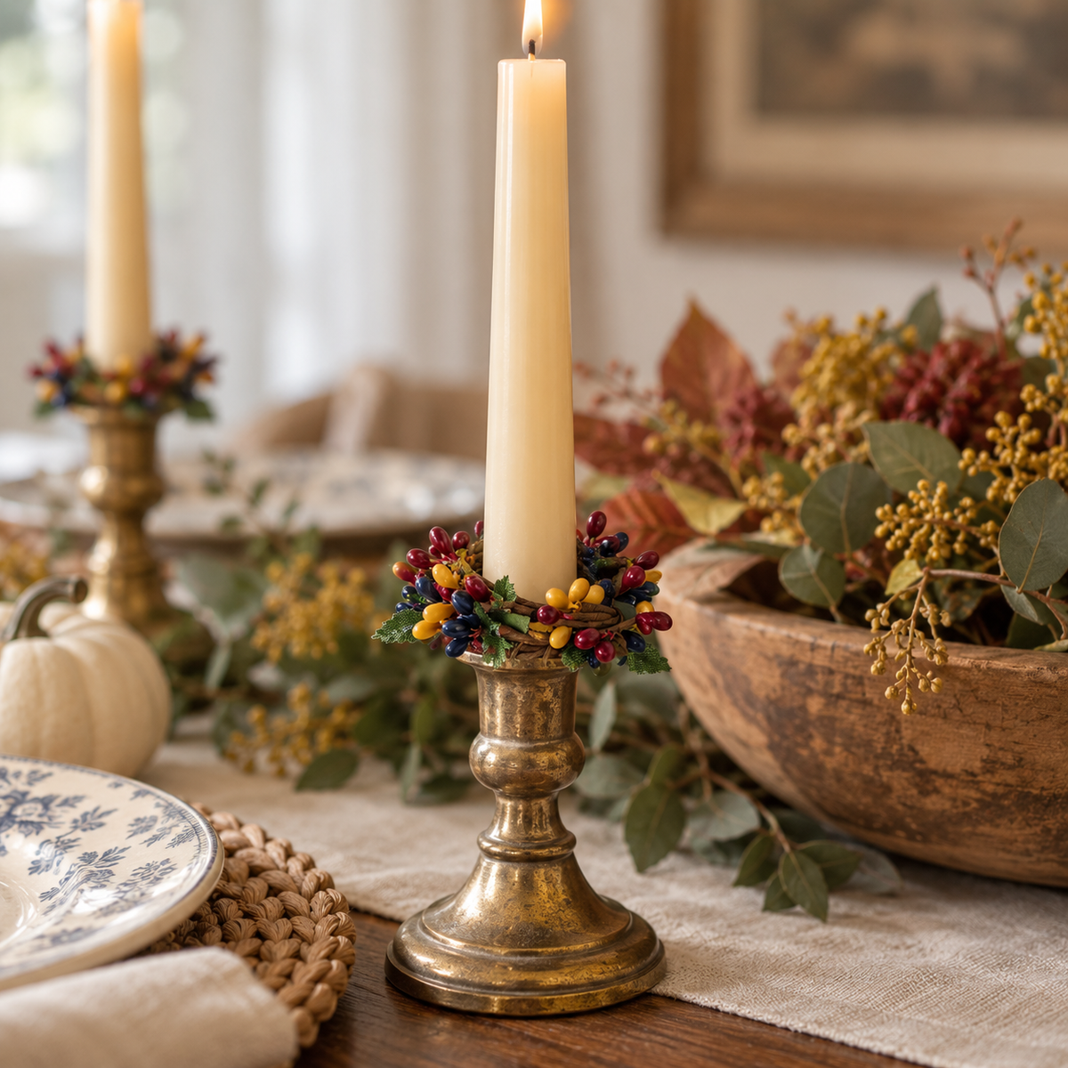 A lit cream pillar candle in a distressed brass candlestick holder with a berry garland, set on a rustic table runner with autumn decorations including a wooden bowl, eucalyptus, and a small pumpkin.