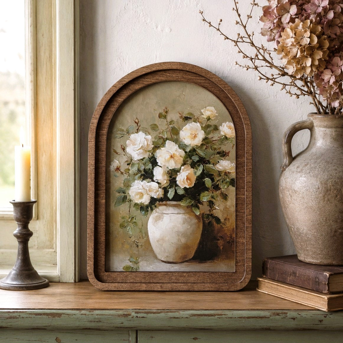 Arched wooden-framed floral wall art leaning on a rustic wooden shelf beside a candle holder and a ceramic vase with dried flowers.