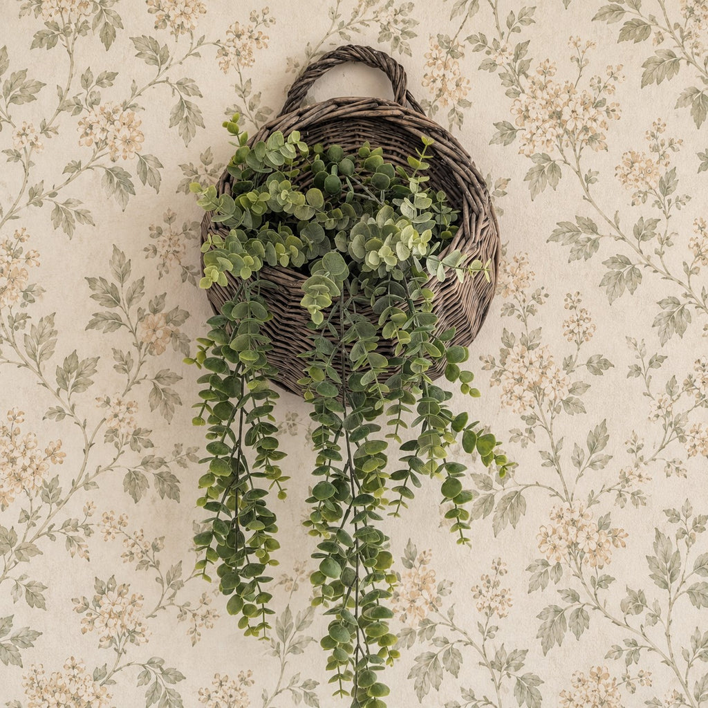 A hanging wicker basket planter with trailing green foliage, displayed against a vintage floral wallpaper background.