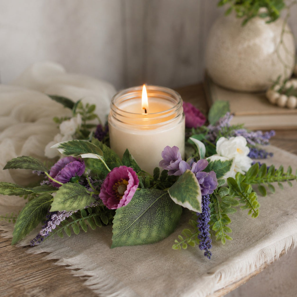 Candle in a glass jar surrounded by purple and white flowers on a textured surface.