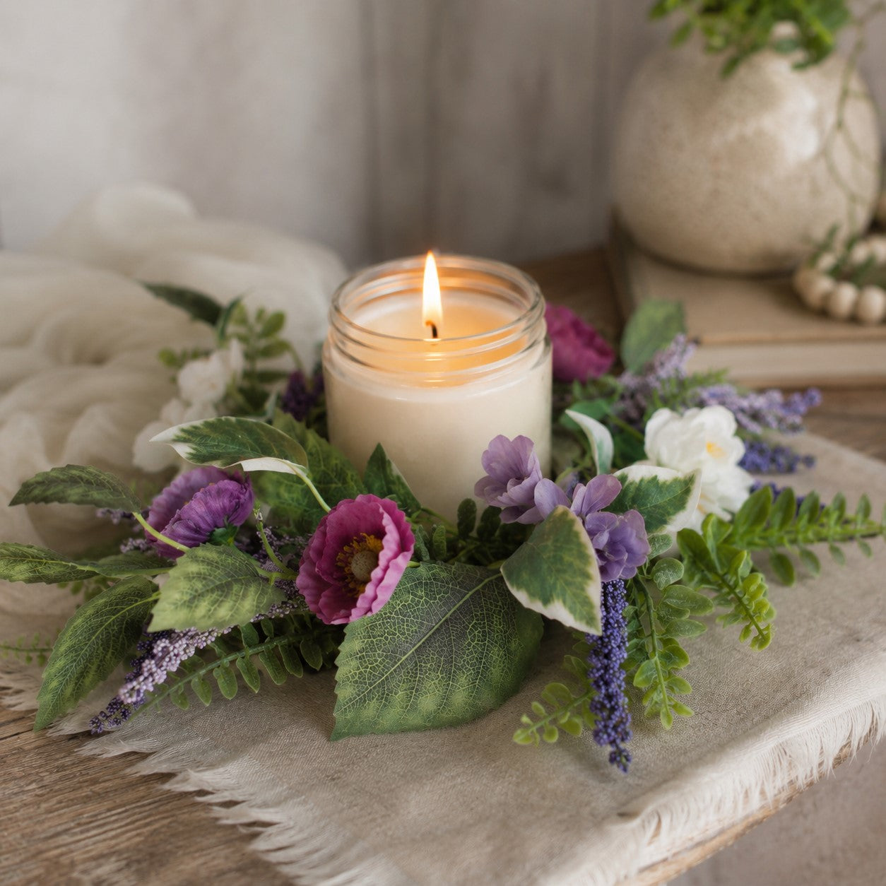 Candle in a glass jar surrounded by purple and white flowers on a textured surface.