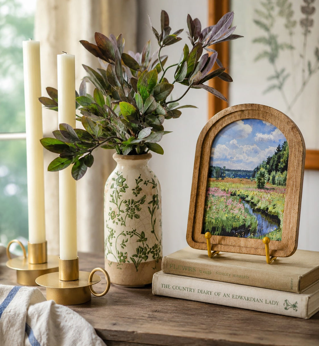 Decorative setup with candles, vase, plant, and framed artwork on a wooden surface.