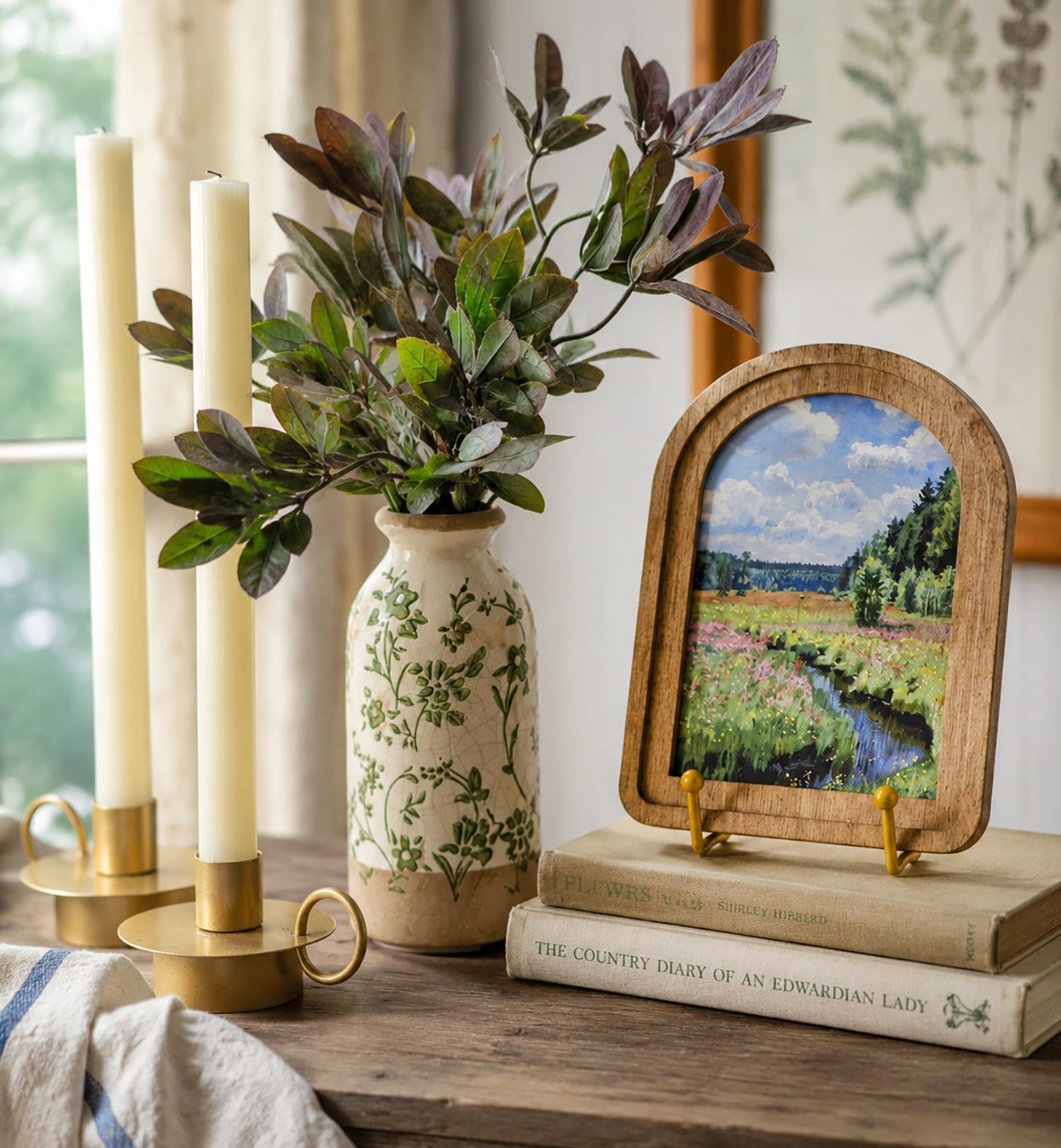 Decorative setup with candles, vase, plant, and framed artwork on a wooden surface.