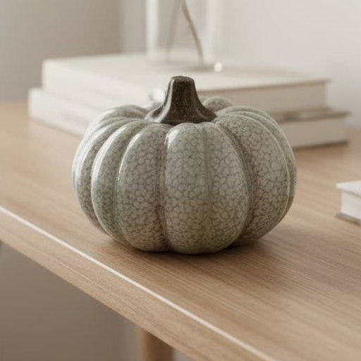 A small ribbed decorative pumpkin in a textured light sage green sits on a light wood console table in a softly lit room, with stacked books and a vase of dried stems behind it.