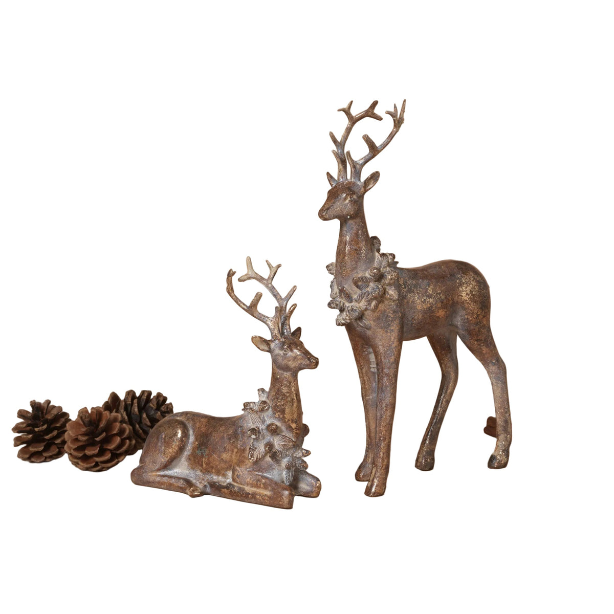 Two rustic metal deer figurines with a distressed bronze finish, one standing and one sitting, styled with pinecones and cinnamon sticks on a warm beige background.