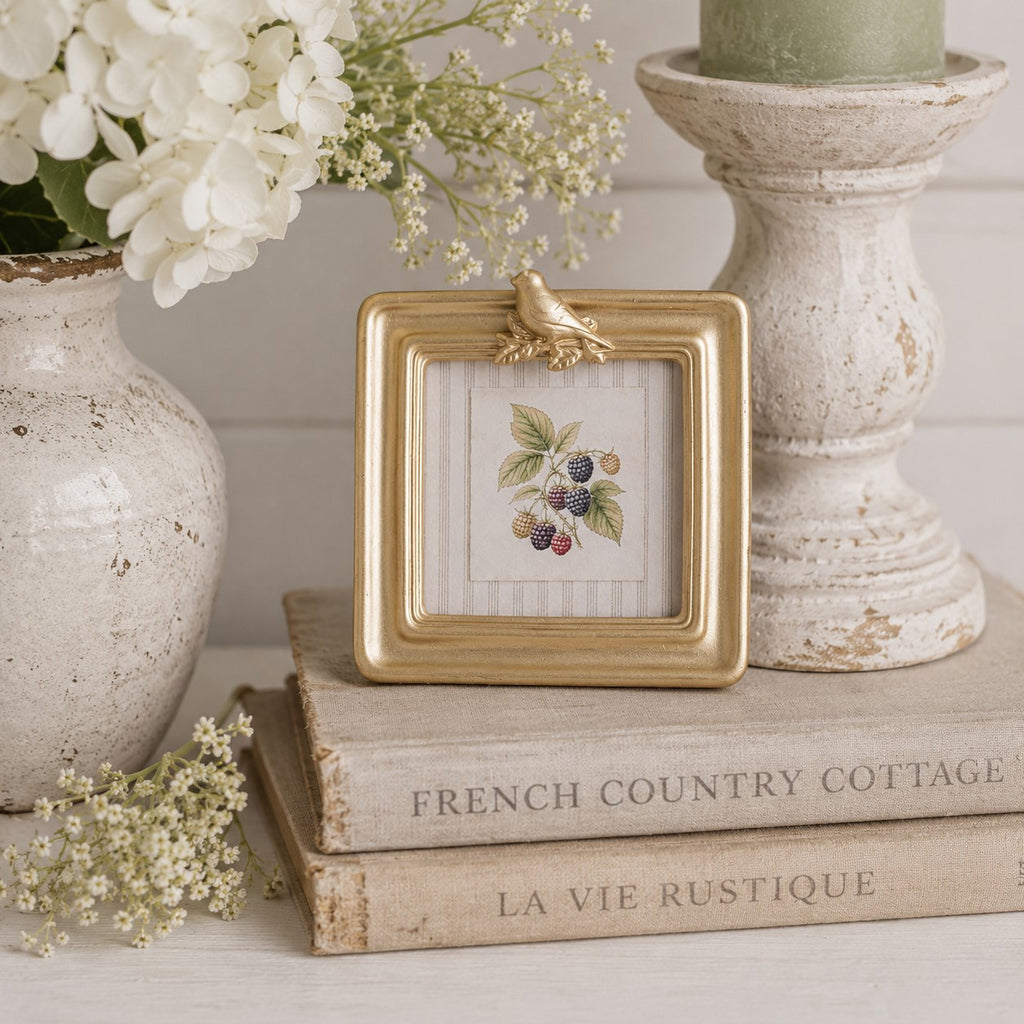 A square gold picture frame with a small bird ornament on the top corner, holding a cross-stitch or printed berry illustration, styled on top of two vintage books beside a vase of white flowers and a distressed white candle holder.