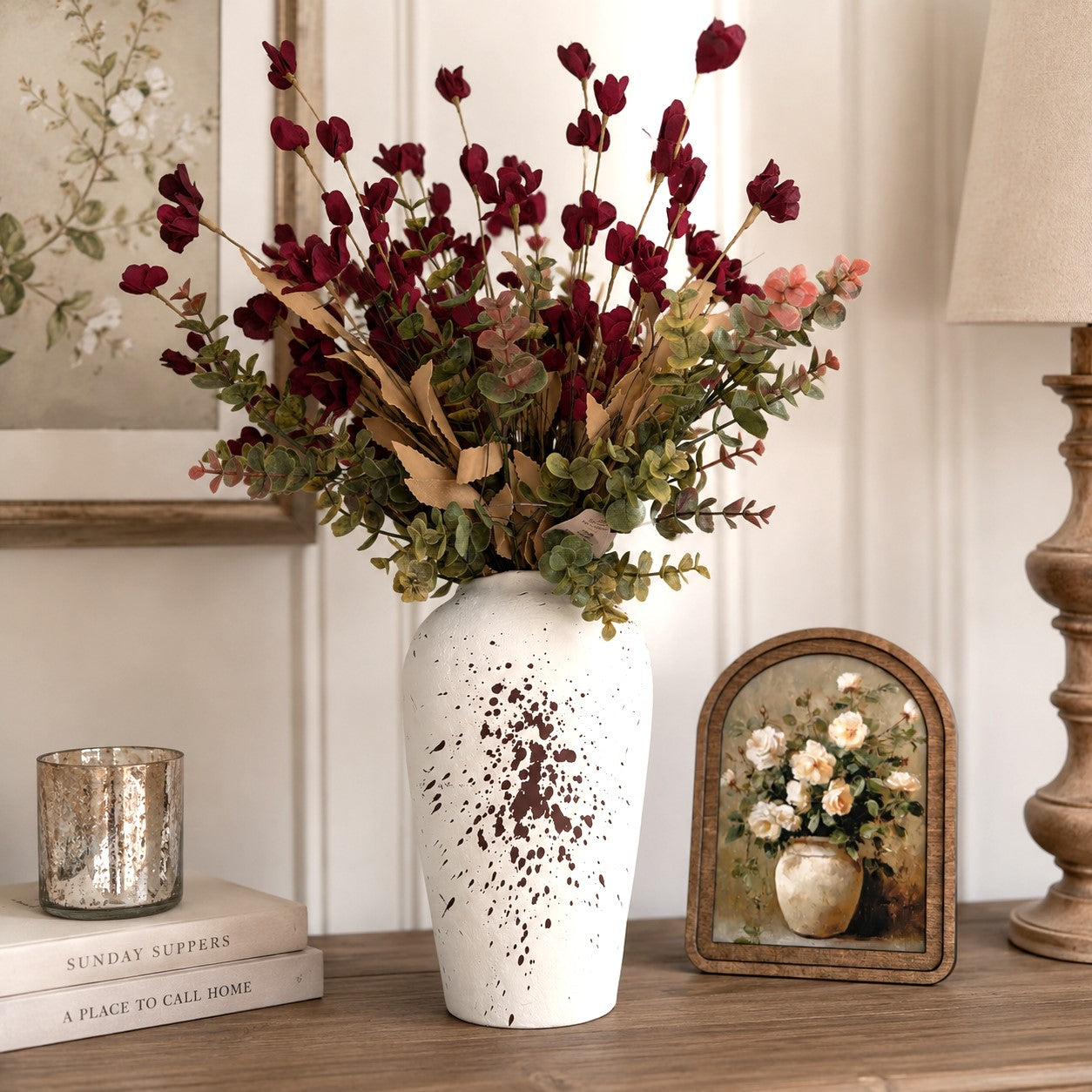A white ceramic vase with brown speckled accents, filled with an artificial floral arrangement of burgundy berries, green eucalyptus, and dried beige stems, displayed on a wooden console table in a styled interior setting.
