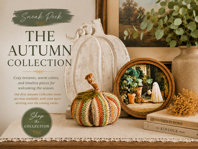 A styled autumn-themed product display with a knitted striped pumpkin in the foreground, a large white pumpkin, a round wooden mirror, books, dried flowers, and a vase with greenery. Text on the left reads 'Sneak Peek THE AUTUMN COLLECTION' with a 'Shop the Collection' badge.