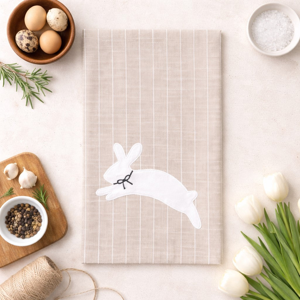 Cream colored kitchen towel featuring hopping rabbit graphic with botanical flowers and greenery in gray tones