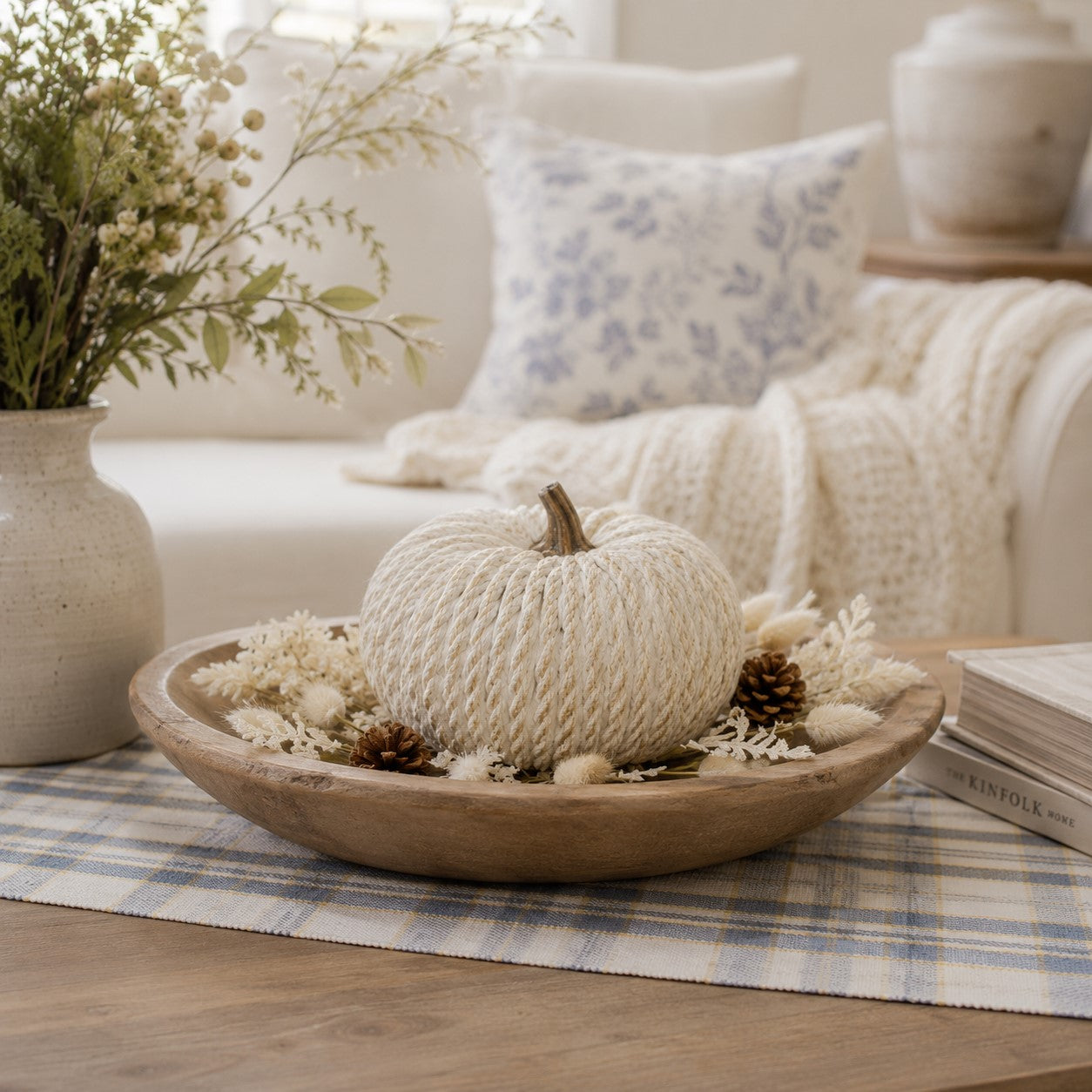 A decorative rope-wrapped pumpkin sits in a wooden bowl with dried flowers and pinecones on a plaid runner in a softly styled living room setting.