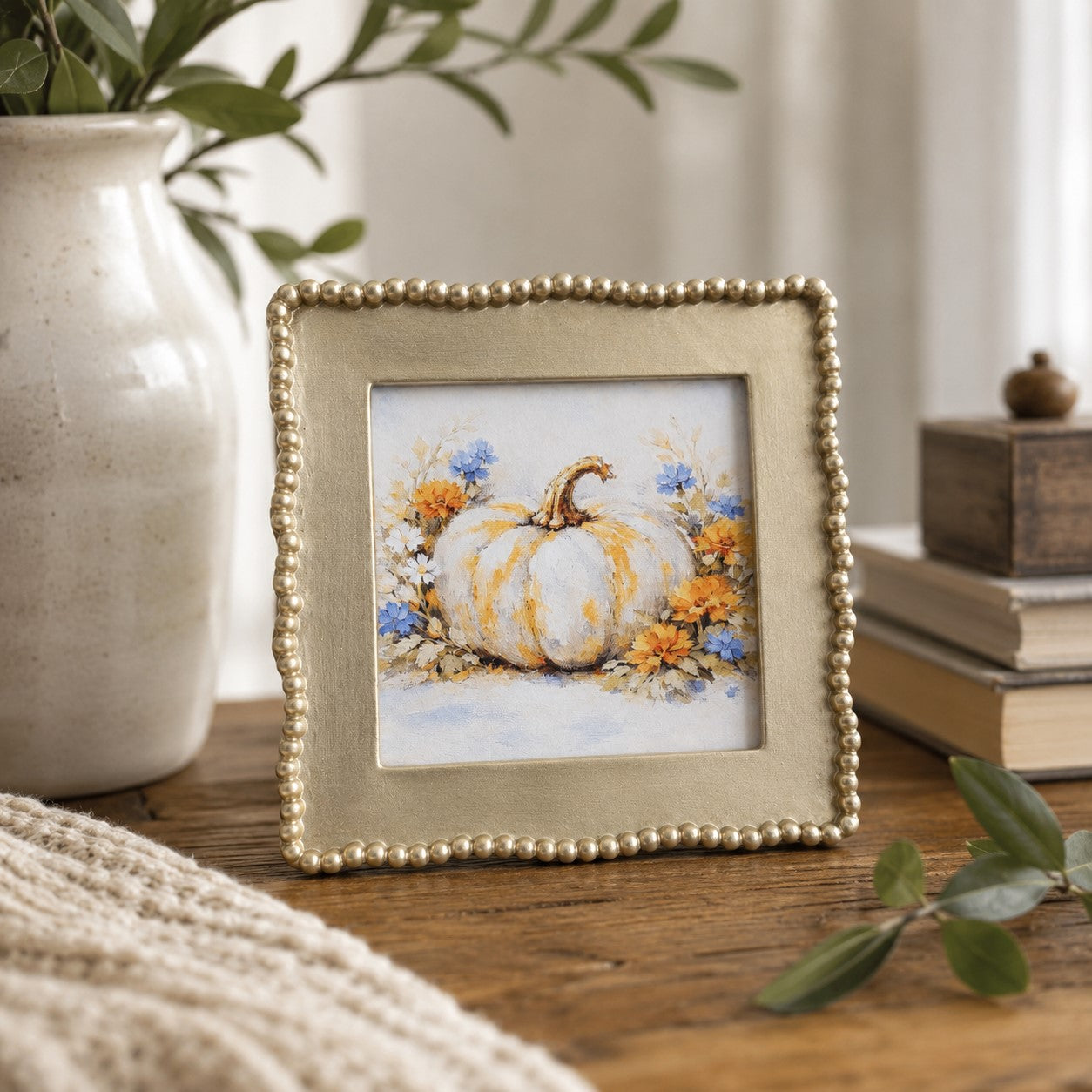 A small square framed art print featuring a painted white pumpkin with orange and blue flowers, styled on a wooden table with a ceramic vase, books, and greenery in a home decor setting.
