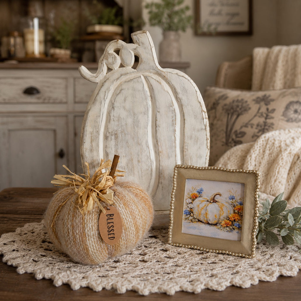 A styled autumn tabletop scene with a large distressed white pumpkin decoration, a smaller beige yarn pumpkin tied with twine and a 'BLESSED' tag, a small framed floral pumpkin print, and a cream crocheted doily on a wooden surface.