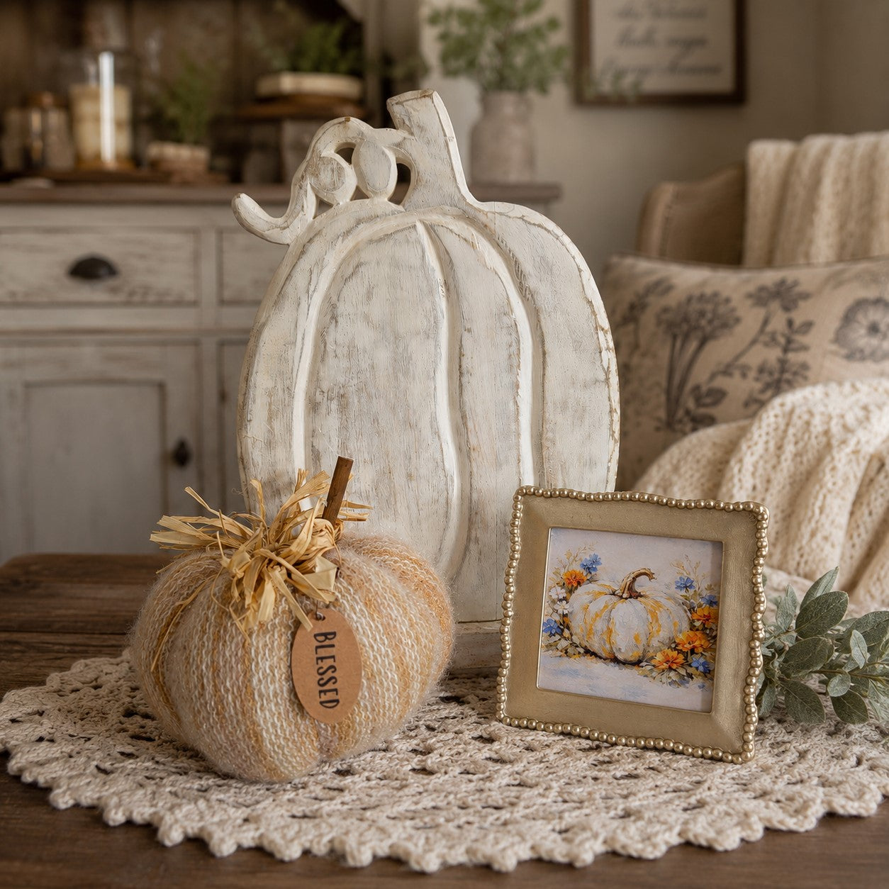 A styled autumn tabletop scene with a large distressed white pumpkin decoration, a smaller beige yarn pumpkin tied with twine and a 'BLESSED' tag, a small framed floral pumpkin print, and a cream crocheted doily on a wooden surface.