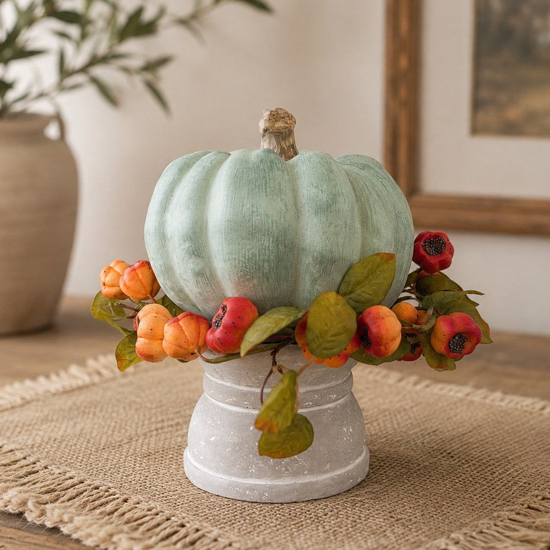 A decorative faux pumpkin centerpiece in a soft mint green color, displayed on a white pedestal stand with an autumn garland of orange berries and green leaves, set on a burlap runner in a home interior.