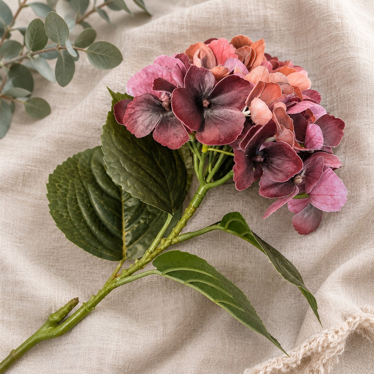 A bouquet of dried or artificial hydrangea stems with deep pink, burgundy, and peach blossoms, green leaves, and a green stem, arranged on a textured beige linen fabric background.