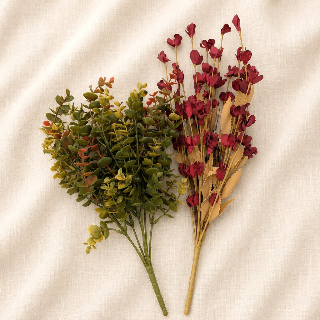 Two artificial floral stems laid flat on a cream fabric background. One stem has small green leaves with hints of yellow and orange, and the other has clusters of dark red flowers with tan leaves.