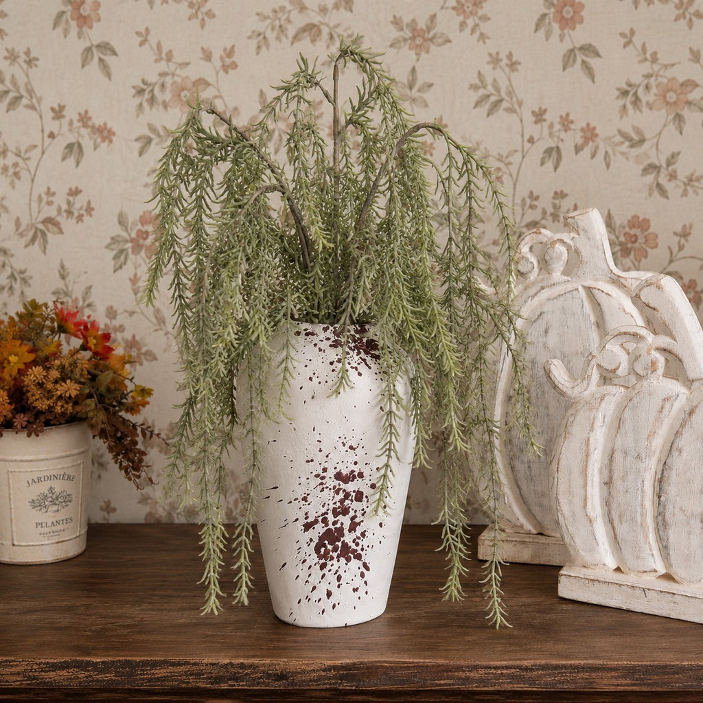 A white speckled ceramic vase holding a green trailing rosemary plant, displayed on a wooden table against a floral wallpaper background, with a white decorative pumpkin and a small potted plant nearby.