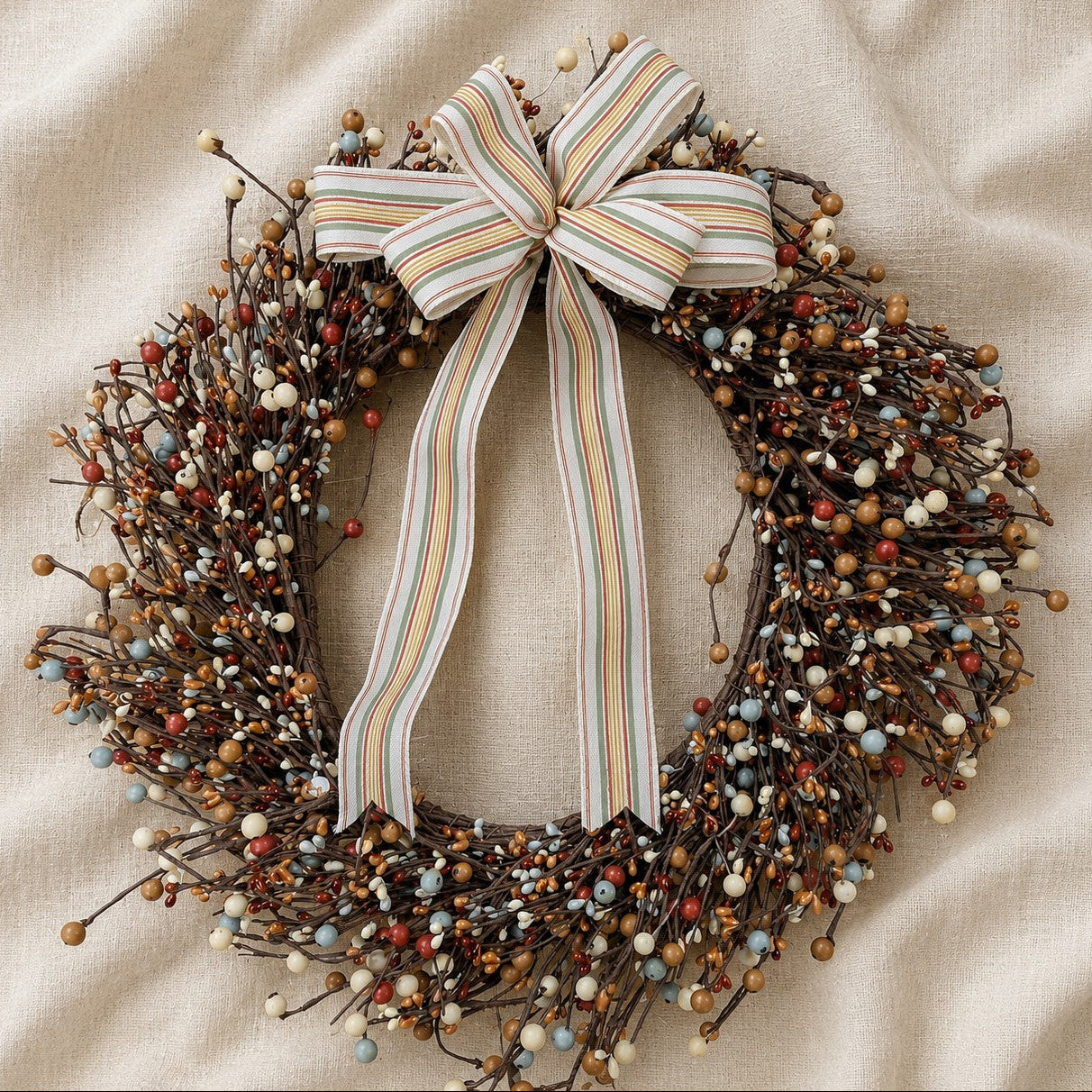 A rustic holiday wreath made of brown branches, decorated with small white, blue, and red berries, and topped with a large cream, beige, and brown striped bow, displayed on a beige fabric background.