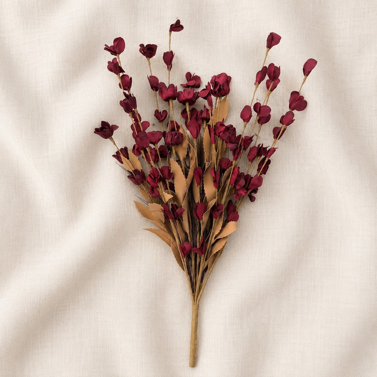 A dried floral stem bundle with deep burgundy petals and tan leaves, arranged on a textured off-white fabric background.