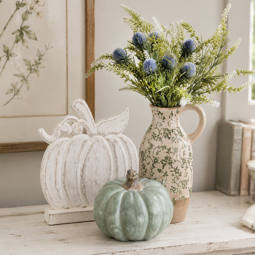 A styled autumn tabletop scene with a small ribbed sage-green pumpkin in front of a floral cream pitcher filled with greenery and blue thistle stems, alongside a distressed white pumpkin decoration.